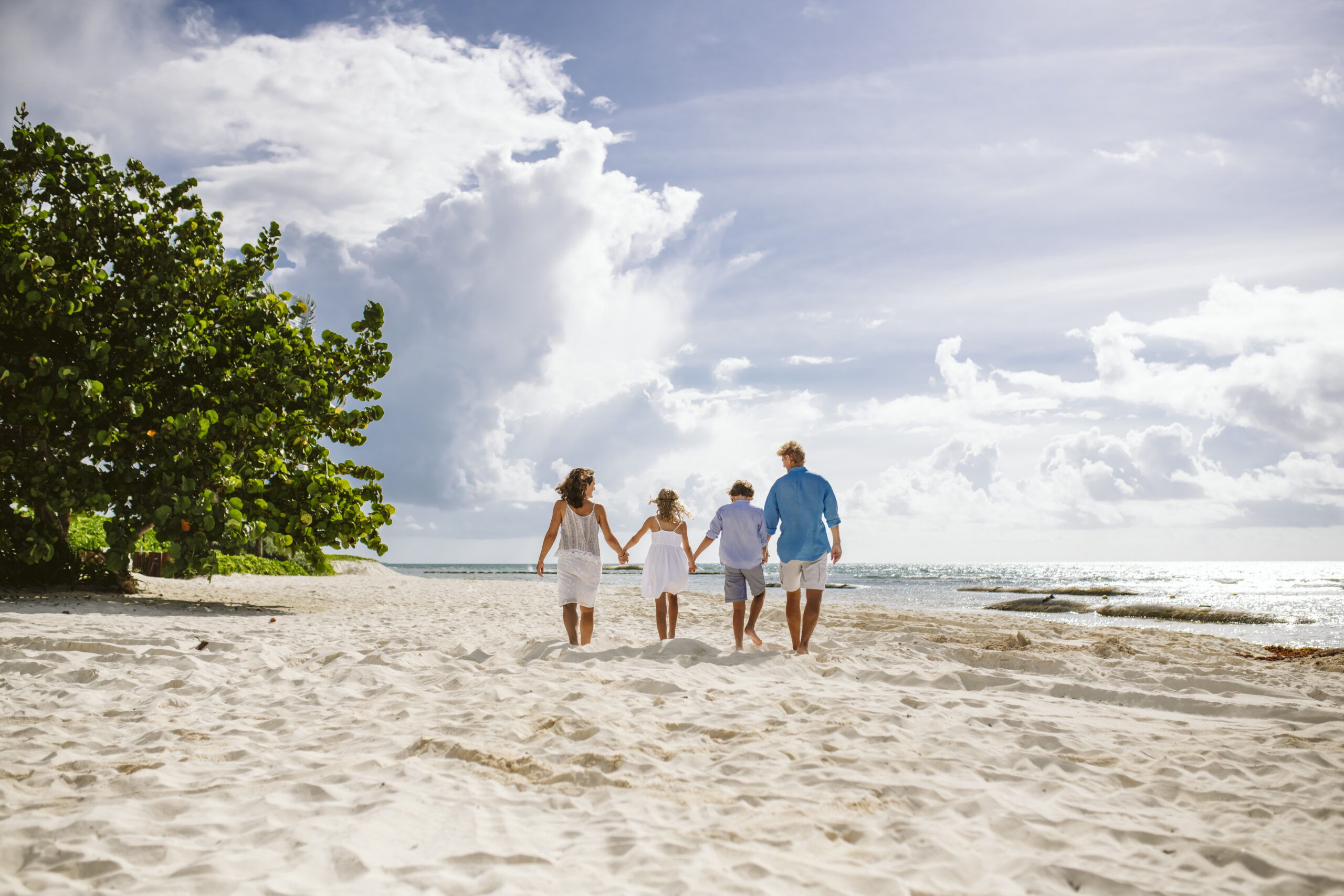 Family of four walking hand in hand on a sandy beach under a blue sky.